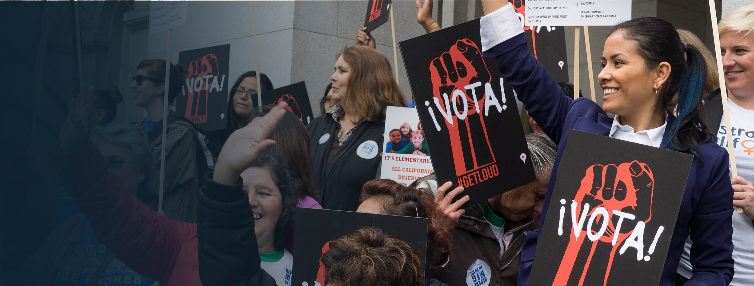 Photo shows a group of women with protest signs that say Vota! with illustrated red power fists on them. The diverse group of women are cheering together.
