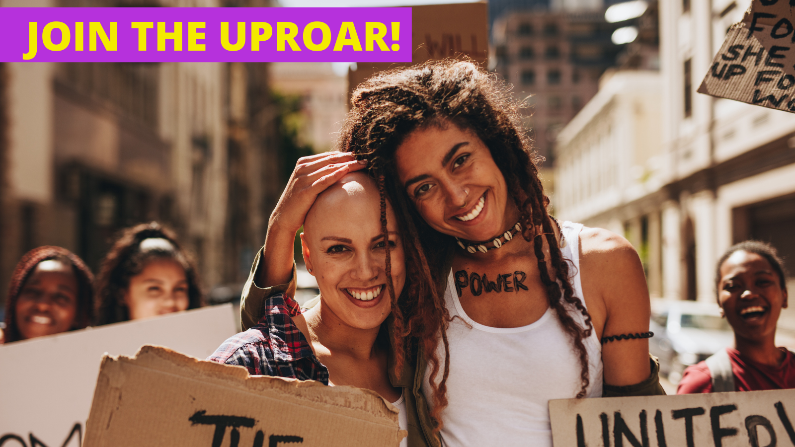 Two women appear side by side and appear to be at a protest. The woman on the left is bald, and lighter skinned. The woman on the right is a person of color with long dreadlocks, she has her hand atop the head of the woman on the left. They are both holding signs that are removed from the frame, but the one on the right appears to say united in all caps.