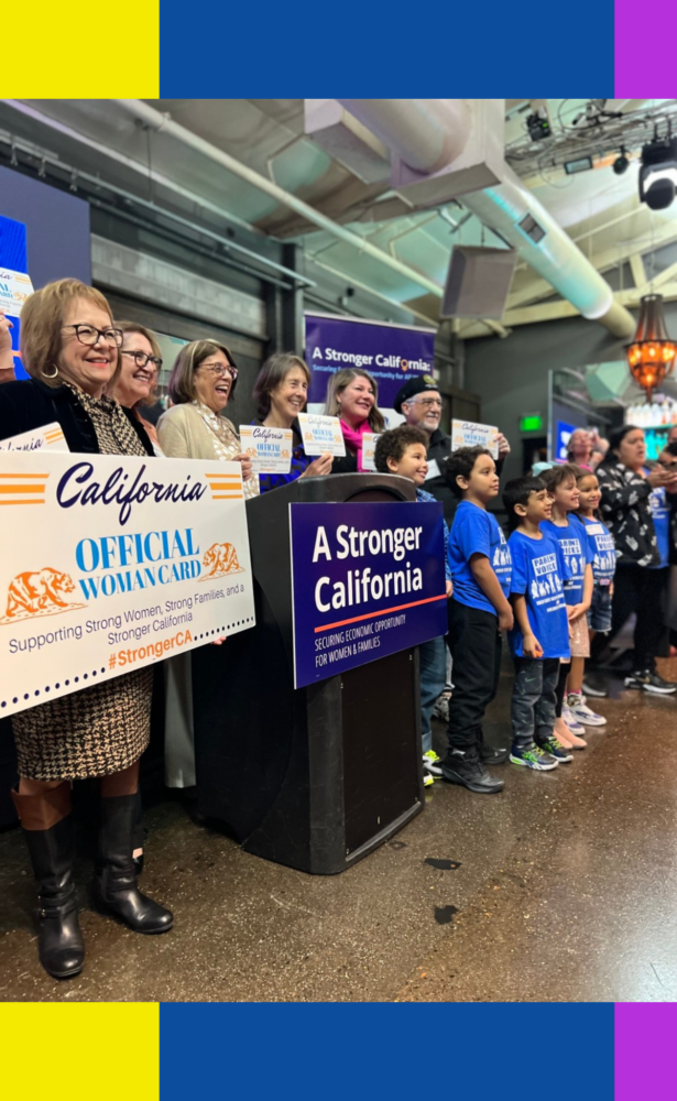 Image of lawmakers, advocates, children, smiling and holding up signs celebrating the Stronger California Advocates Network. They are lined up, this photo was taken to the left of them while they smile at another camera.