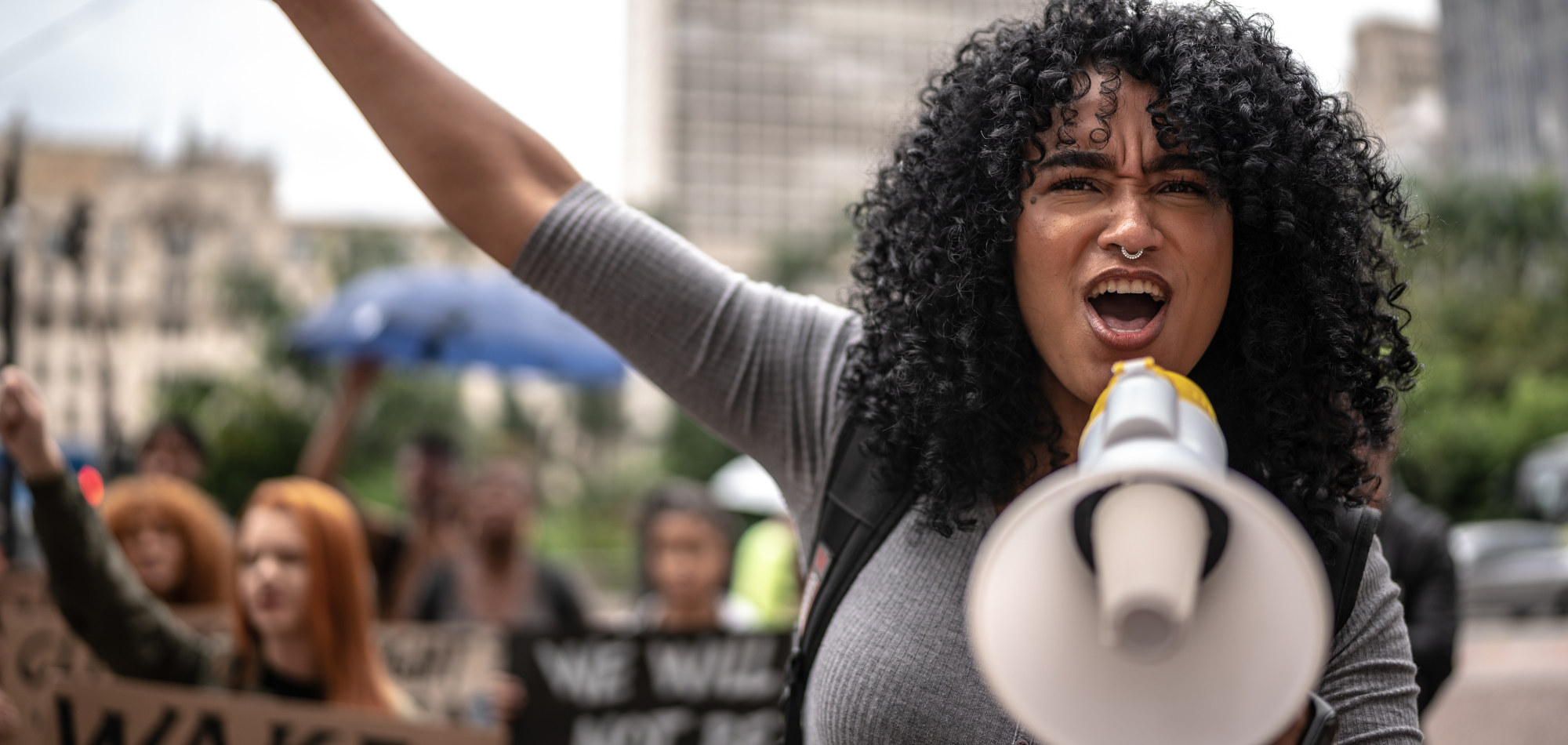 A young femme adult is shouting into a white and yellow megaphone. She is wearing a gray T-shirt and has dark, curly, shoulder-length hair. She looks determined and inspired. Behind her are other protesters with signs. They're marching in an urban setting.