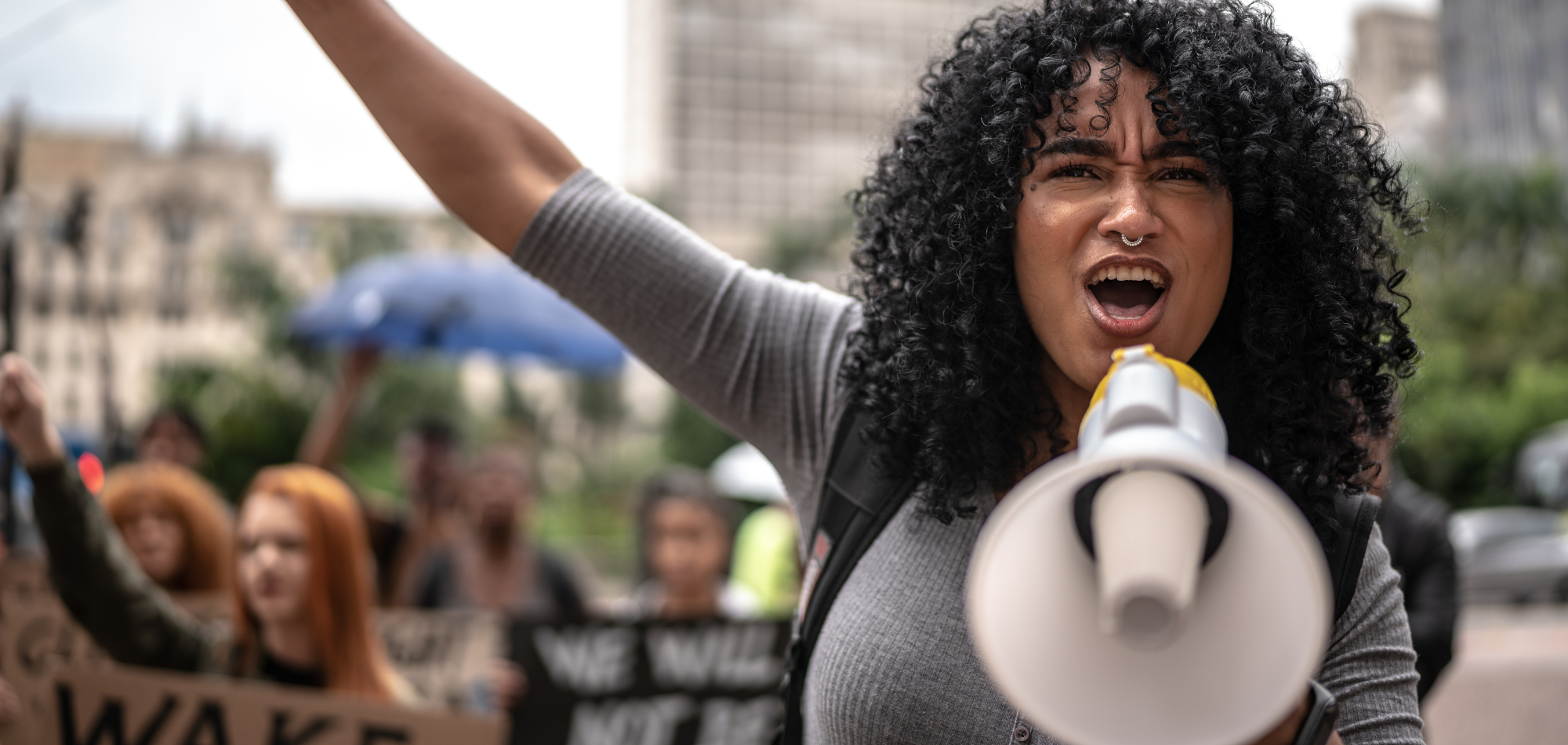 A young femme adult is shouting into a white and yellow megaphone. She is wearing a gray T-shirt and has dark, curly, shoulder-length hair. She looks determined and inspired. Behind her are other protesters with signs. They're marching in an urban setting.