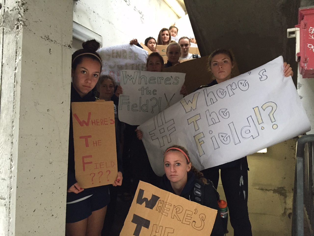 A photo of a group of young women Berkeley field hockey players in sweatshirts, looking into the camera with a challenge in their eyes, holding signs that say WTF, Where's The Field?