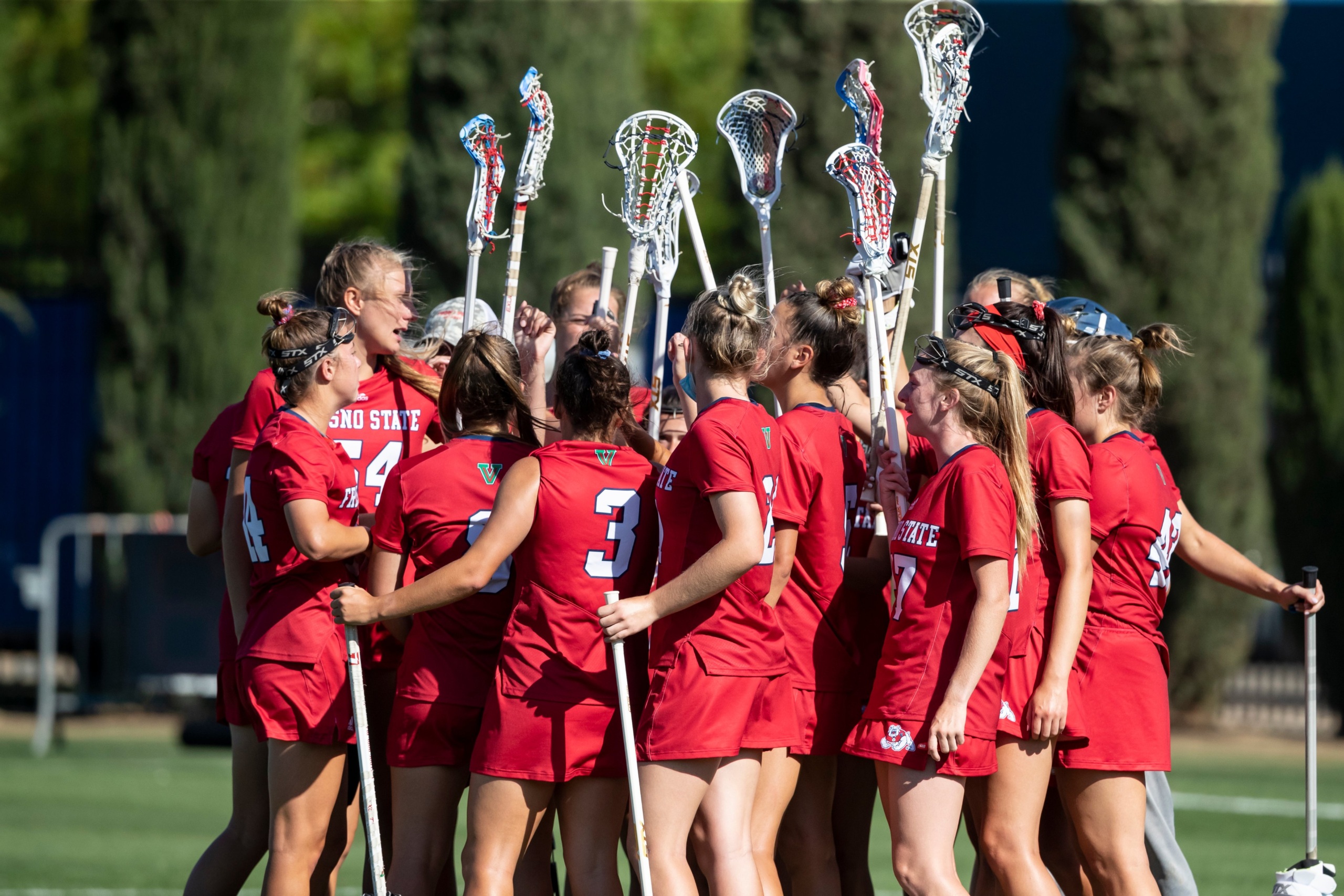 A group of about 15 femme student lacrosse players huddle in a circle, wearing red Fresno State sports uniforms, facing away from the camera, lifting their lacrosse sticks into the air in victory.