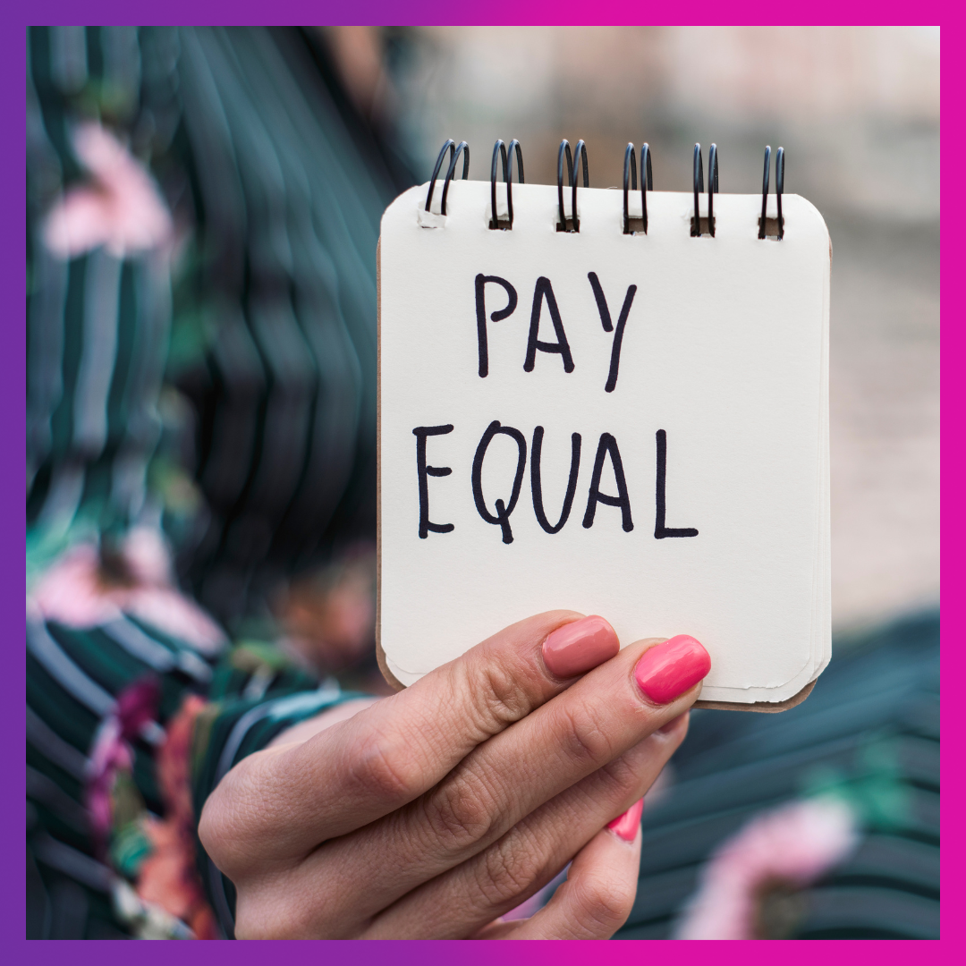 Photo of a hand with painted nails holding a notepad with text written on it that says Pay Equal