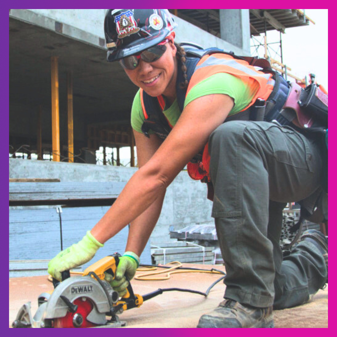 Photo of a woman construction worker wearing a hardhat, safety glasses, a toolbelt, and an orange vest using a circular saw on a large sheet of plywood