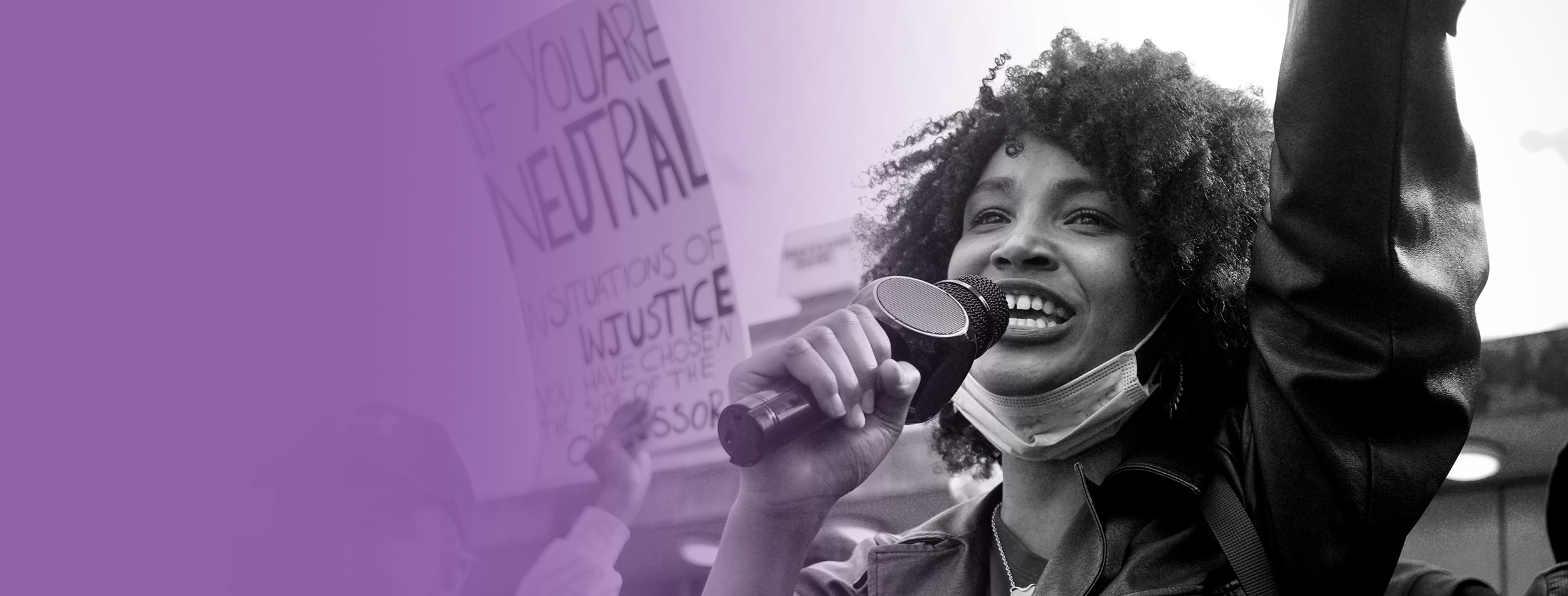 Black and white photo of a protester speaking into a microphone and raising their fist in the air.